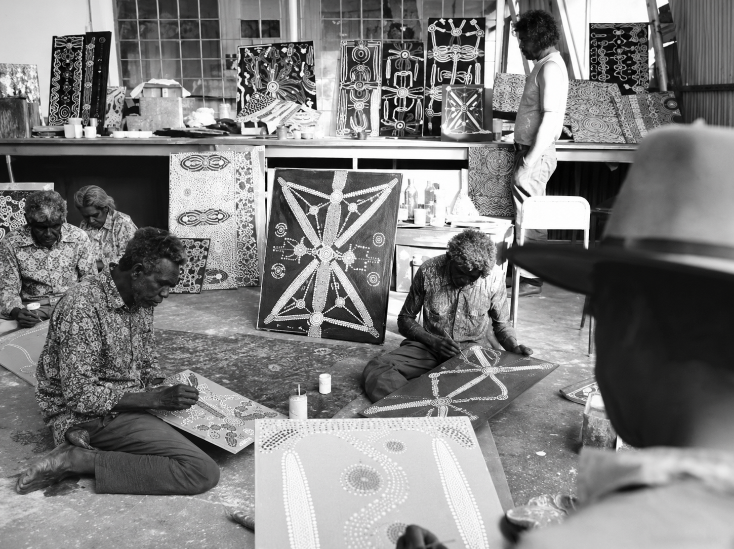 Historic black-and-white photograph of Aboriginal artists painting early Western Desert Art canvases inside the men's painting shed