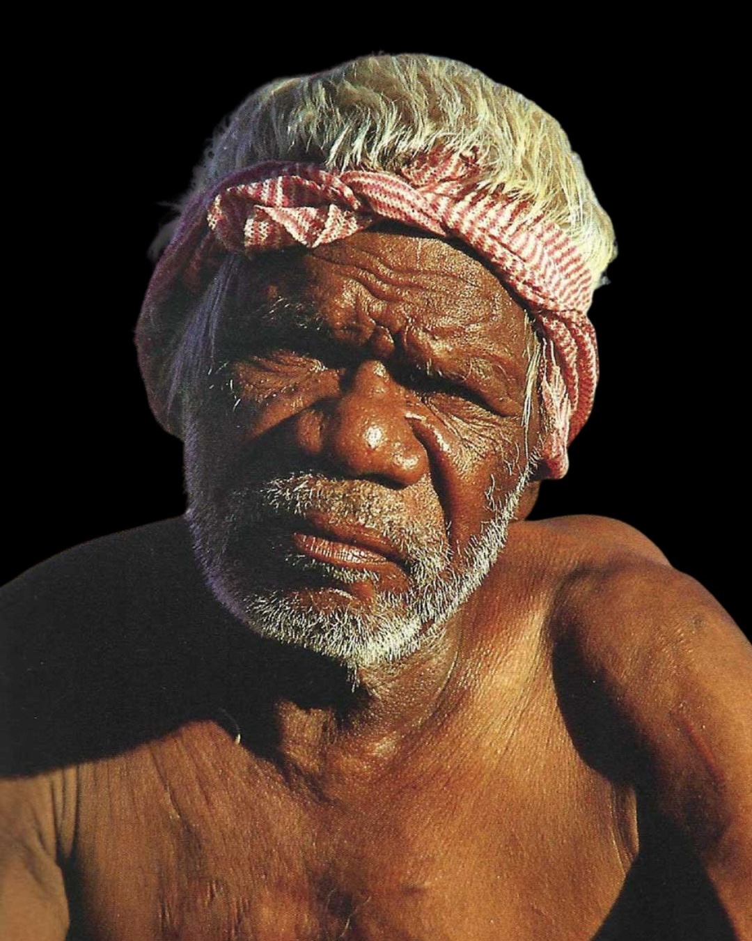 Portrait photograph of East Kimberley Aboriginal artist Rover Thomas wearing a red headband against a dark background
