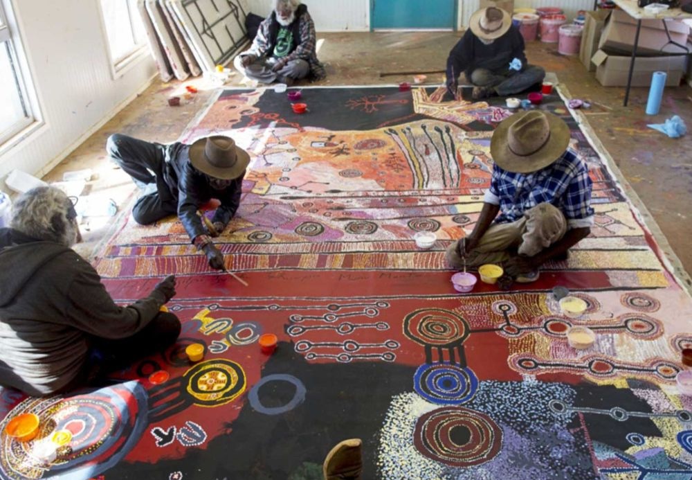 Anangu artists working together on a large contemporary APY Lands painting, applying traditional and contemporary Western Desert designs inspired by Country and Tjukurpa.