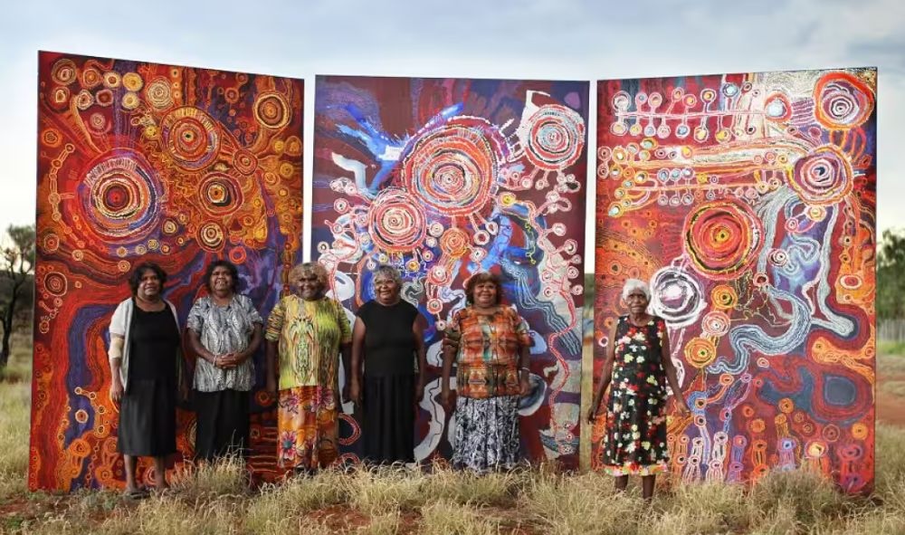 Anangu women artists standing beside a large-scale APY Lands collaborative artwork depicting the Seven Sisters Dreaming through vibrant colour, concentric circles, and ancestral pathways.