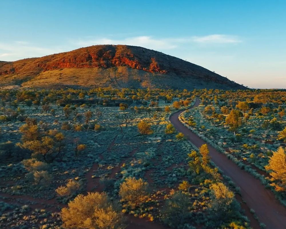 Desert landscape in the Ngaanyatjarra Lands showing a rocky red range, spinifex plains, and winding road through remote Western Australia.