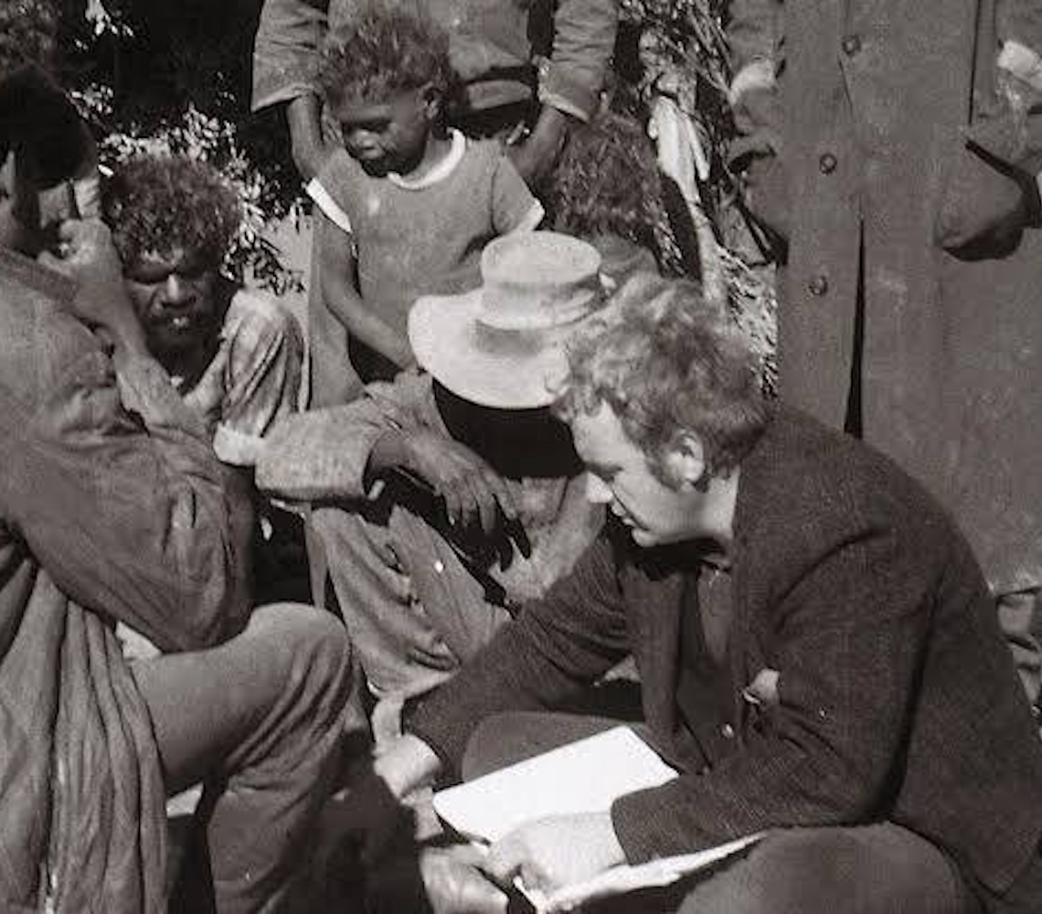 Historic black and white photograph of Geoffrey Bardon speaking with Aboriginal men at Papunya during the early development of Western Desert painting