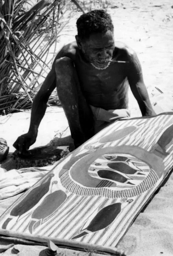 Aboriginal artist painting a bark painting using natural ochre pigments on a prepared sheet of eucalyptus bark in Arnhem Land.
