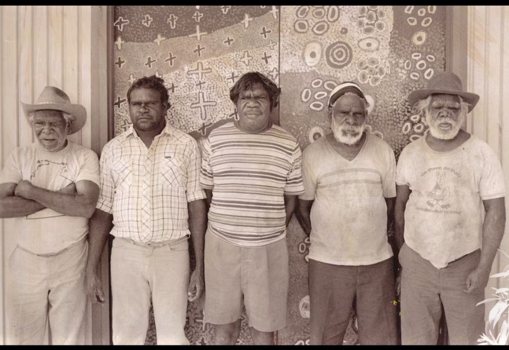 Group portrait of Warlpiri men standing before painted ceremonial designs at Yuendumu, Central Australia, representing the custodians of Warlpiri language, law, ceremony, and artistic traditions.