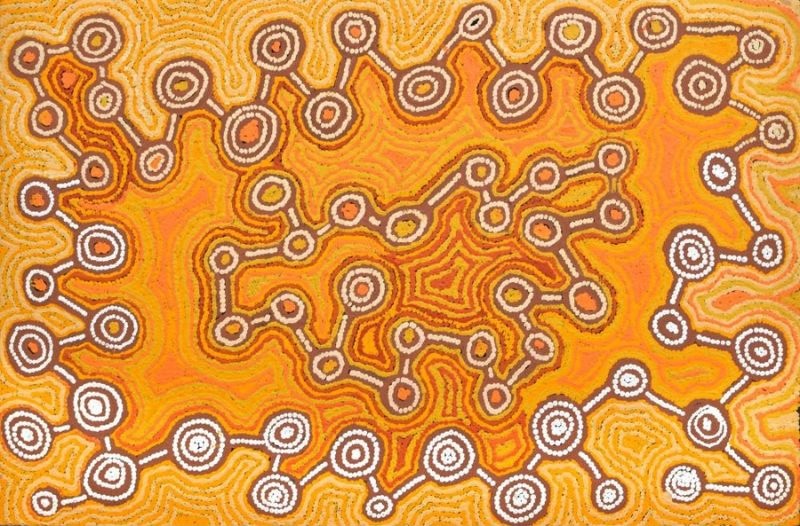 A horizontal Aboriginal painting glowing with warm desert colours of golden yellow, bright orange, ochre, cream, chocolate brown and white. The centre is dominated by a large radiant orange landform with softly flowing contours that spreads across the painting like a glowing island. Around its edges, dozens of concentric circles of different sizes are linked by dark brown pathways, creating a chain of interconnected ceremonial sites. Smaller angular motifs sit within the orange field, while thousands of tiny cream and white dots build every line and shape. The contrast between the bright orange centre and the pale yellow background creates a strong sense of depth and movement, giving the impression of an aerial view of Country alive with connected places and travelling routes.