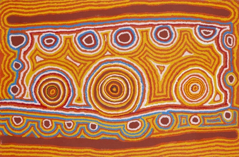 A bright horizontal Aboriginal painting dominated by vivid golden yellow, burnt orange, deep red-brown, sky blue and white. Three large concentric circles stretch across the centre, the middle circle noticeably larger than the others, with each ring built from finely dotted bands of yellow, orange, brown and white. Smaller circles line the upper and lower edges, linked by pale blue pathways that weave around the composition. Angular stepped motifs and triangular forms fill the spaces between the circles, while broad horizontal bands frame the top and bottom. Thousands of small dots create flowing contour-like lines that give the painting a rhythmic, almost vibrating appearance, evoking interconnected ceremonial sites across the Western Desert.