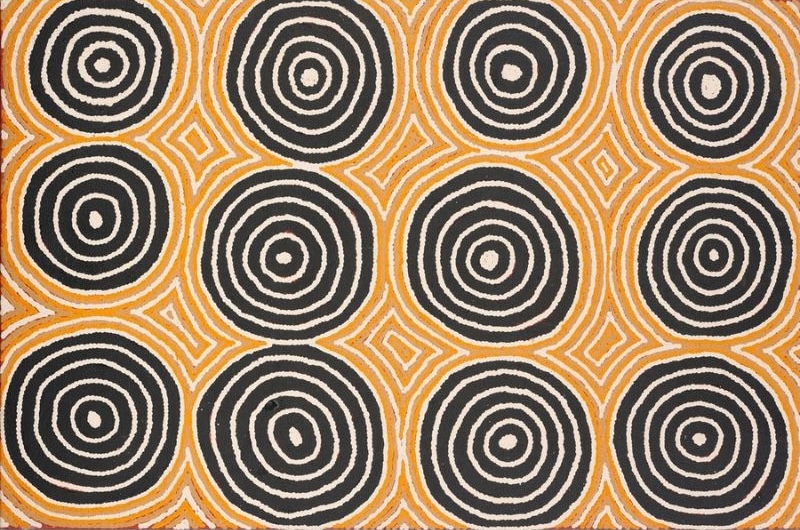 Untitled by Warlimpirrnga Tjapaltjarri, 2000. A vertical painting in black, white and golden ochre. Twelve large concentric circles dominate the composition, arranged in a neat grid across a warm ochre background. Fine white and ochre lines radiate between the circles, forming small diamond-shaped patterns that create a balanced, rhythmic design resembling an aerial map of connected ceremonial sites.