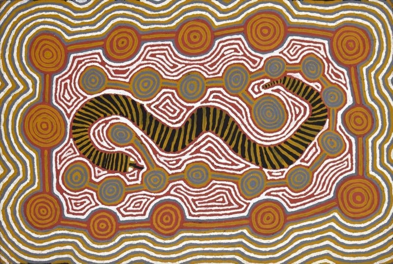 Wanampi Dreaming at Wilkinkarra by Warlimpirrnga Tjapaltjarri, 1996. A horizontal painting in earthy ochres, deep black, white, grey-blue and rusty red. A large black snake with yellow stripes winds across the centre of the composition, surrounded by many concentric circles connected by pathways. Thick undulating bands frame the painting, while intricate dotted lines and geometric patterns create a rhythmic, ceremonial map of the desert landscape.