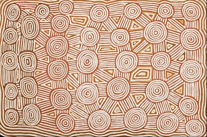 Untitled by Warlimpirrnga Tjapaltjarri, 1999. A horizontal painting in warm ochre, cream and soft brown. Dozens of concentric circles of varying sizes are arranged across the canvas, linked by straight parallel lines and interspersed with triangular and oval motifs. The orderly geometric pattern resembles an aerial map of interconnected ceremonial sites, while the fine dotted outlines create a gentle shimmering effect.