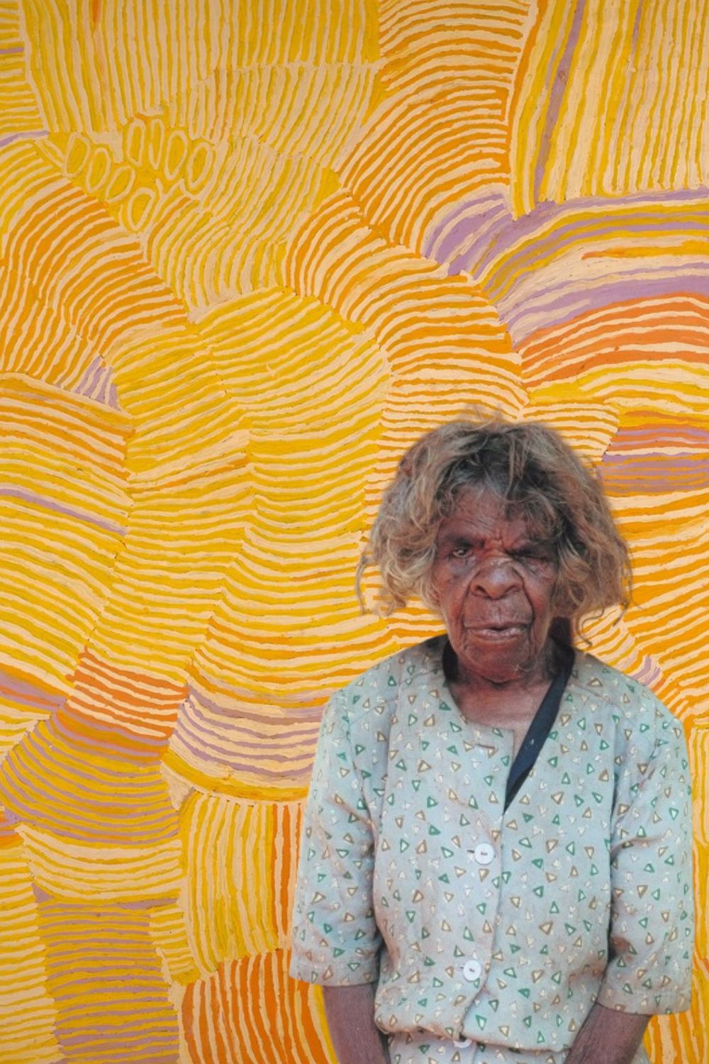 Makinti Napanangka standing before one of her large abstract paintings inspired by Pintupi women's ceremony and ceremonial hair-string traditions.
