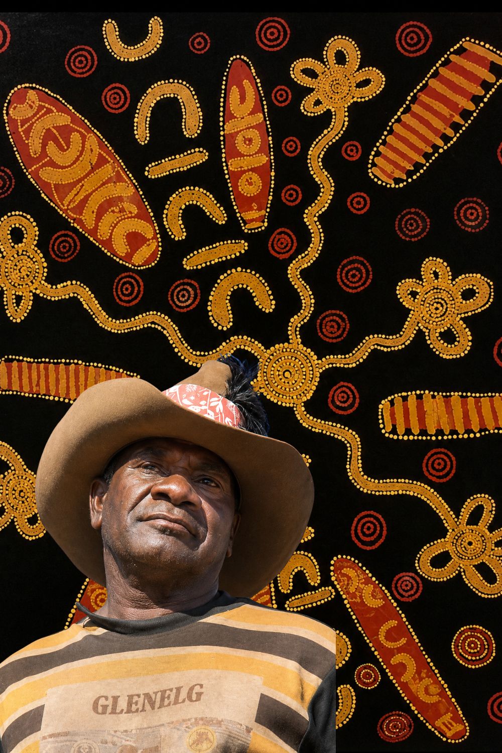 Portrait of Tommy Lowry Tjapaltjarri in front of one of his early Papunya Board paintings featuring Snake Dreaming and sweeping ancestral pathways.