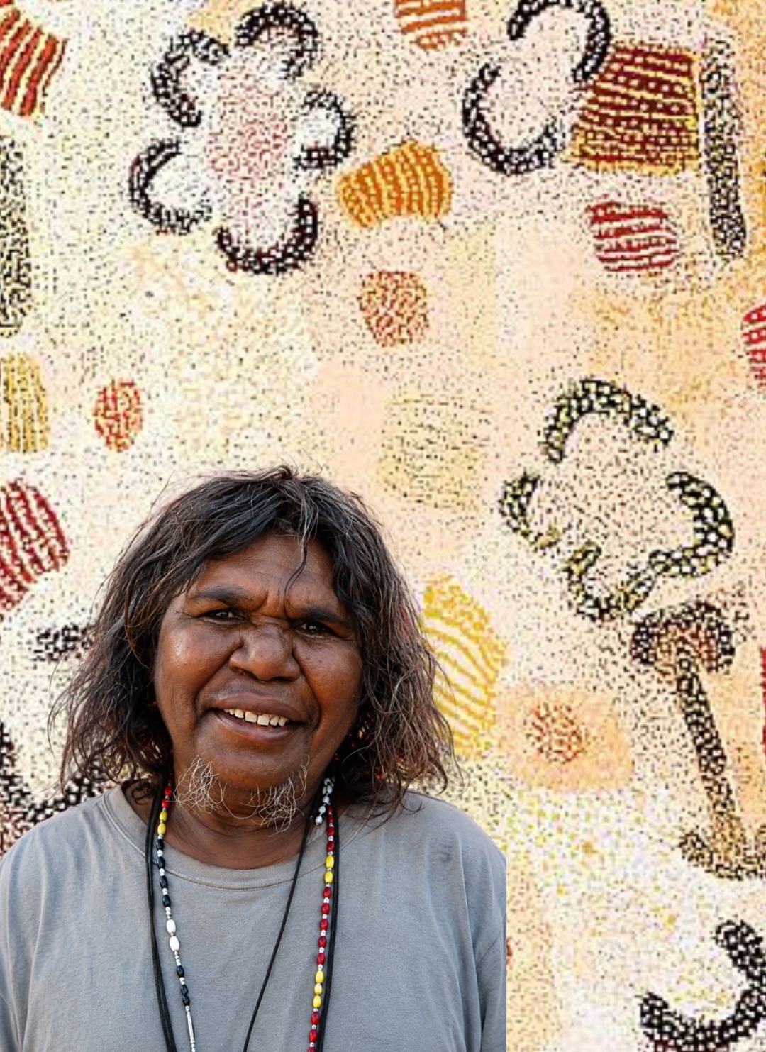 Portrait of Elizabeth Nyumi (c.1947–2019), leading Balgo and Western Desert artist, standing before her Parwalla painting depicting the spinifex, bush foods and sandhills of her father's Country in the Great Sandy Desert.