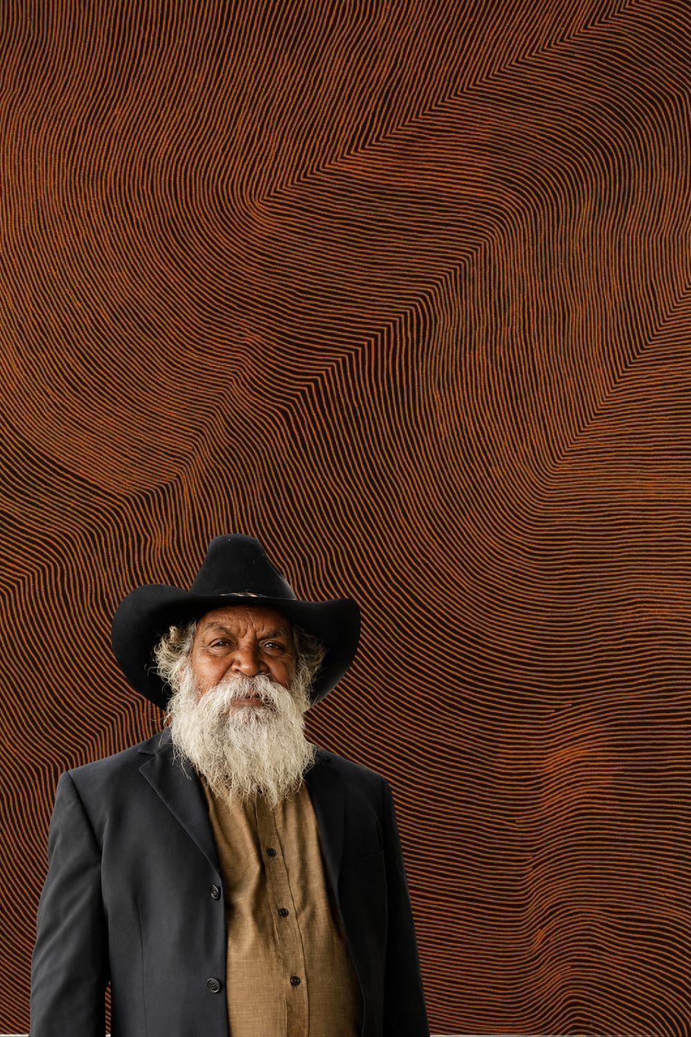Portrait of Aboriginal artist Warlimpirrnga Tjapaltjarri standing before one of his large abstract paintings, featuring thousands of fine parallel ochre and black lines that create shimmering geometric patterns inspired by Tingari Country.