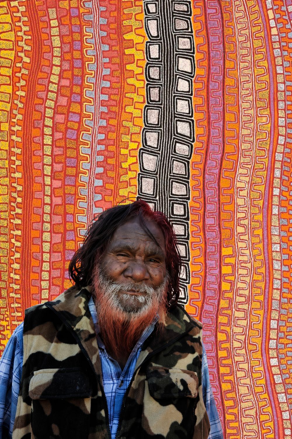 Portrait of Western Desert Aboriginal artist Patrick Tjungurrayi standing in front of a brightly painted vehicle featuring his distinctive geometric desert designs.