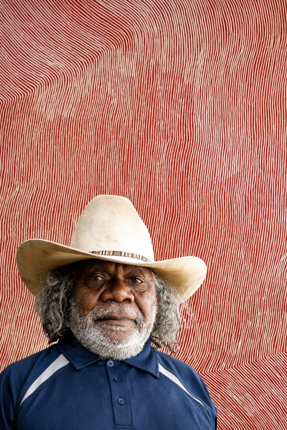 Portrait of George Tjungurrayi standing before one of his red and white Tingari paintings with shimmering parallel lines.