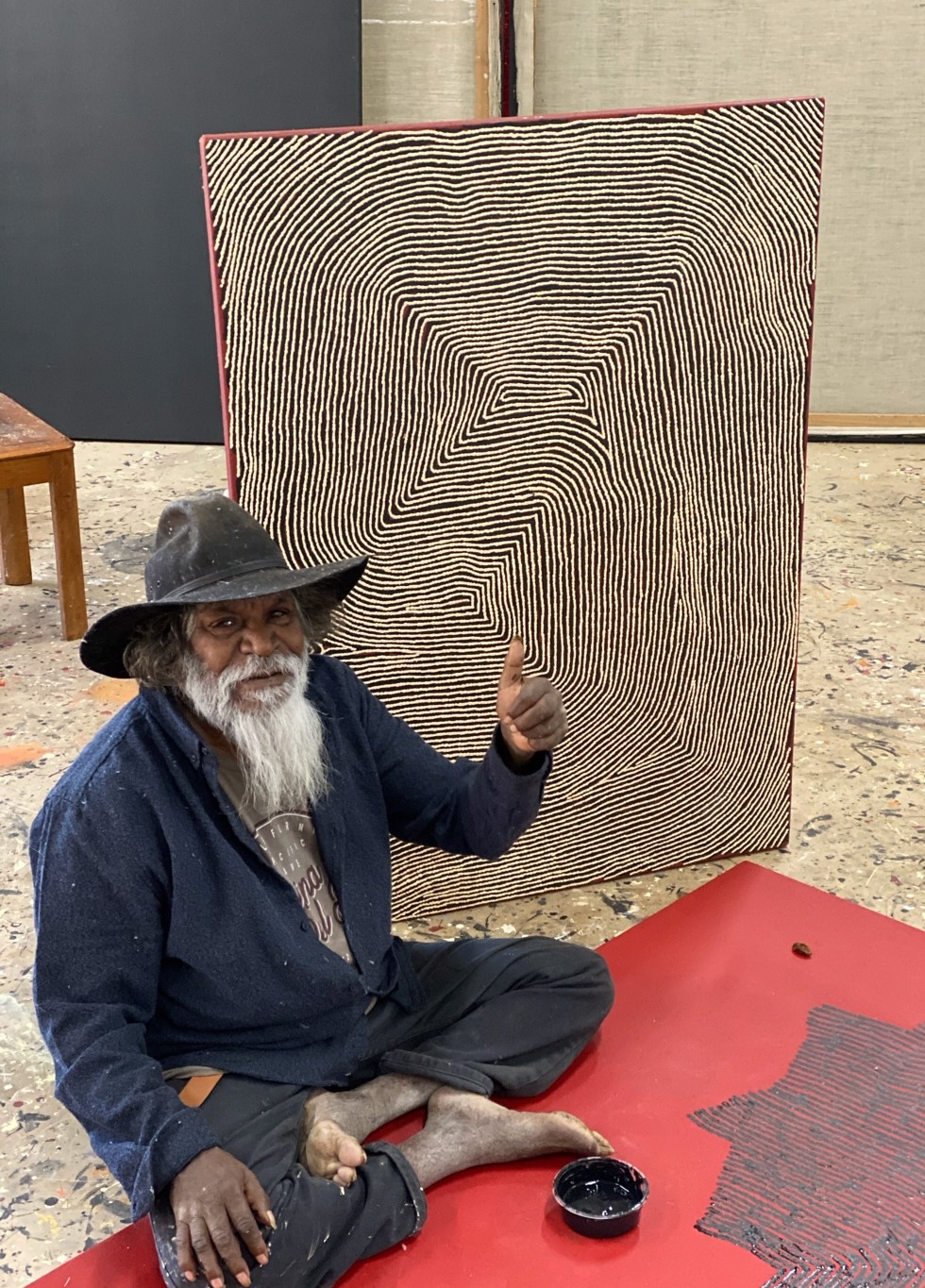 Warlimpirrnga Tjapaltjarri seated barefoot in his studio beside a large monochrome painting of fine parallel white lines on a black background, giving a thumbs-up while working on a new canvas.