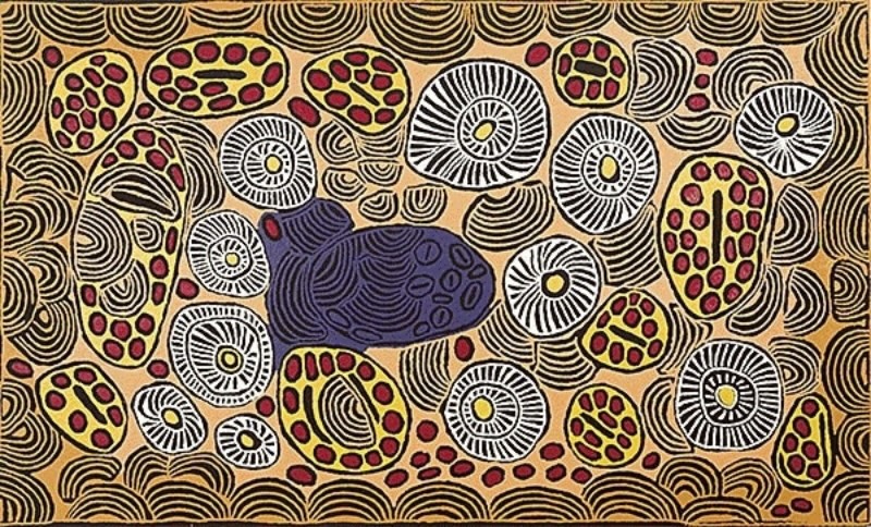 Large ochre, black, white, red and deep blue Aboriginal painting by Ningura Napurrula. The surface is densely patterned with black concentric arcs, large white circular forms filled with radiating black lines, and irregular yellow shapes containing clusters of red dots. A prominent dark blue oval form filled with flowing lines and smaller oval marks occupies the centre-left.