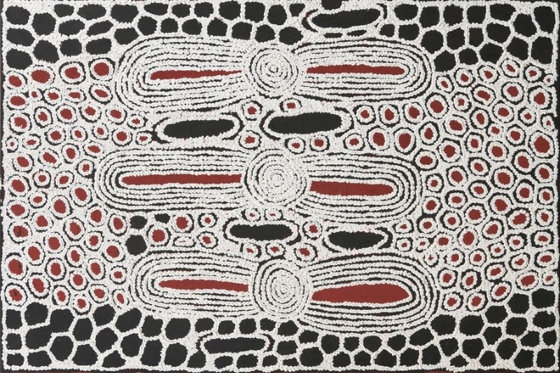 Black, white and red Aboriginal painting by Ningura Napurrula with three concentric circles arranged vertically through the centre. Each circle joins long horizontal oval forms outlined by dense white lines and containing narrow red centres. Hundreds of smaller red circles outlined in white fill the surrounding field, while irregular black shapes enclosed by white outlines form a network-like pattern around the outer edges.
