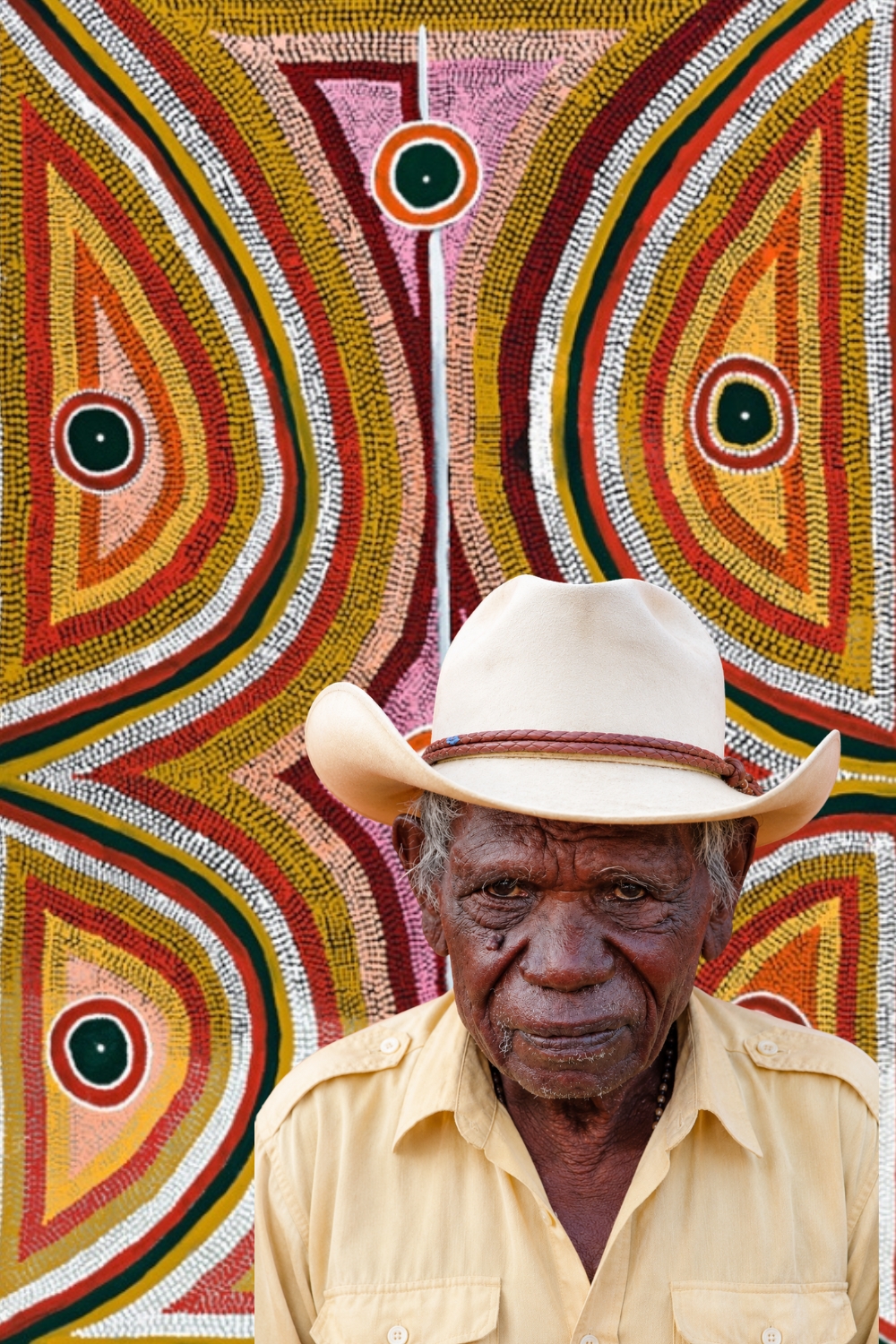 Boxer Milner Portrait of Aboriginal artist Elizabeth Nyumi Nungurrayi standing in front of one of her pale dotted paintings of Parwalla Country.
