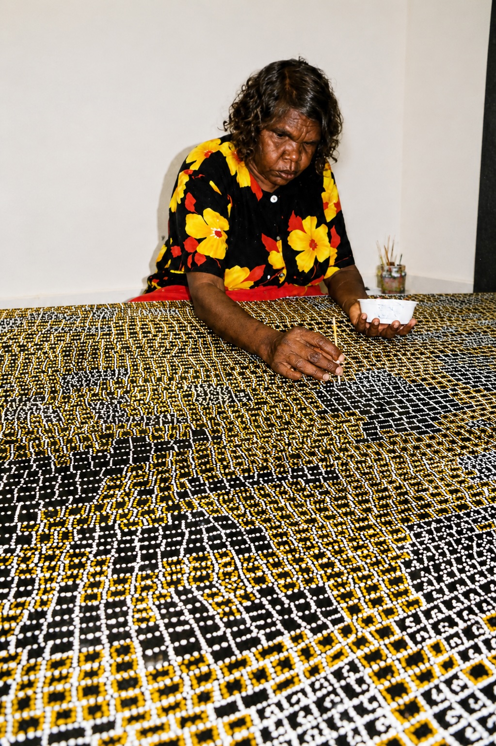 Dorothy Napangardi painting one of her iconic Mina Mina artworks using fine white and yellow dots on a large black canvas.