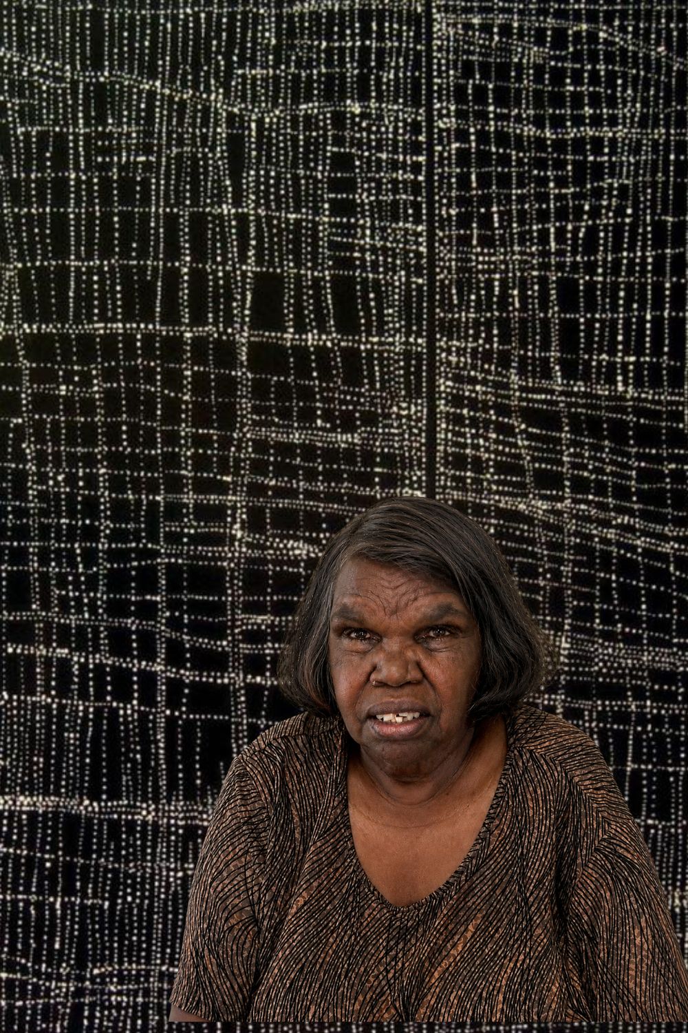 Dorothy Napangardi Portrait | Warlpiri Artist of Mina Mina Portrait of Dorothy Napangardi, celebrated Warlpiri artist and one of the leading painters of the Western Desert art movement, pictured in front of her distinctive black-and-white Mina Mina painting.