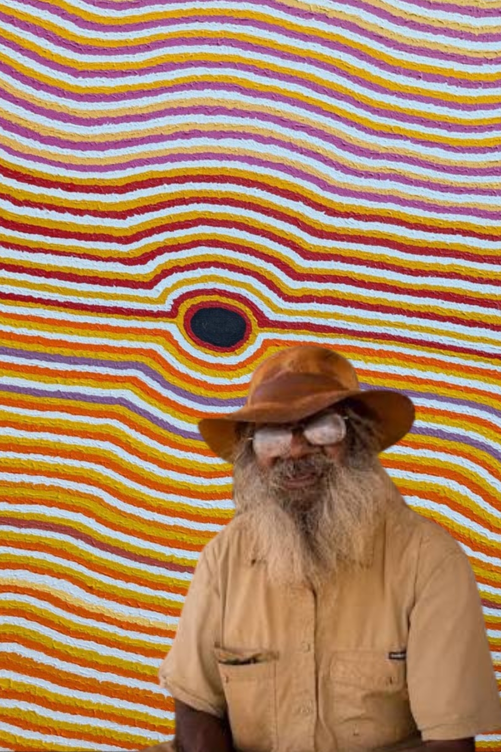 Helicopter Tjungurrayi Portrait of Aboriginal artist Eubena Nampitjin standing before her vibrant Kinyu (Spirit Dog) painting from the Balgo art movement.