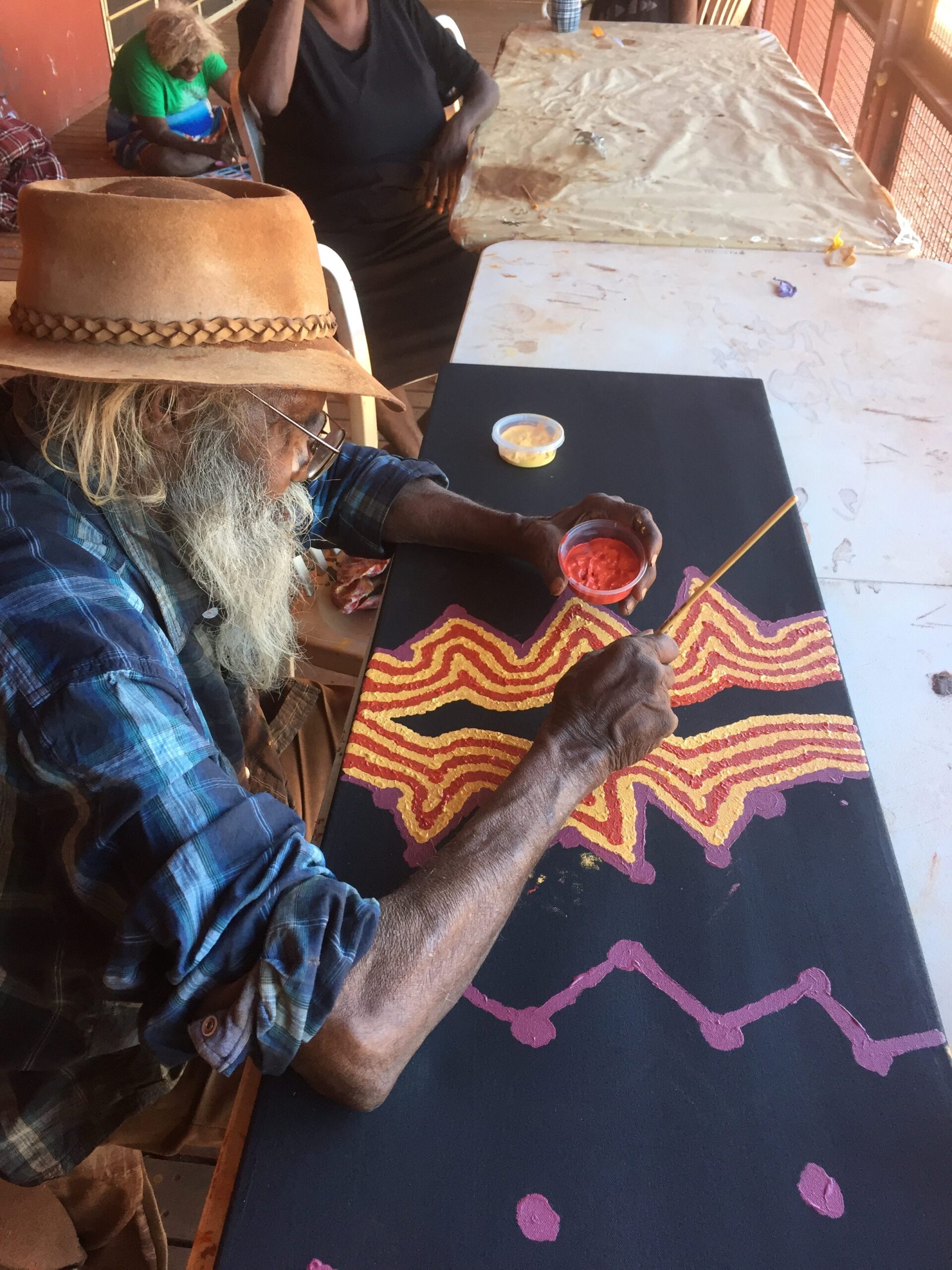 Senior Aboriginal artist Helicopter Tjungurrayi painting parallel red and yellow lines on a black canvas at Balgo, Western Australia.