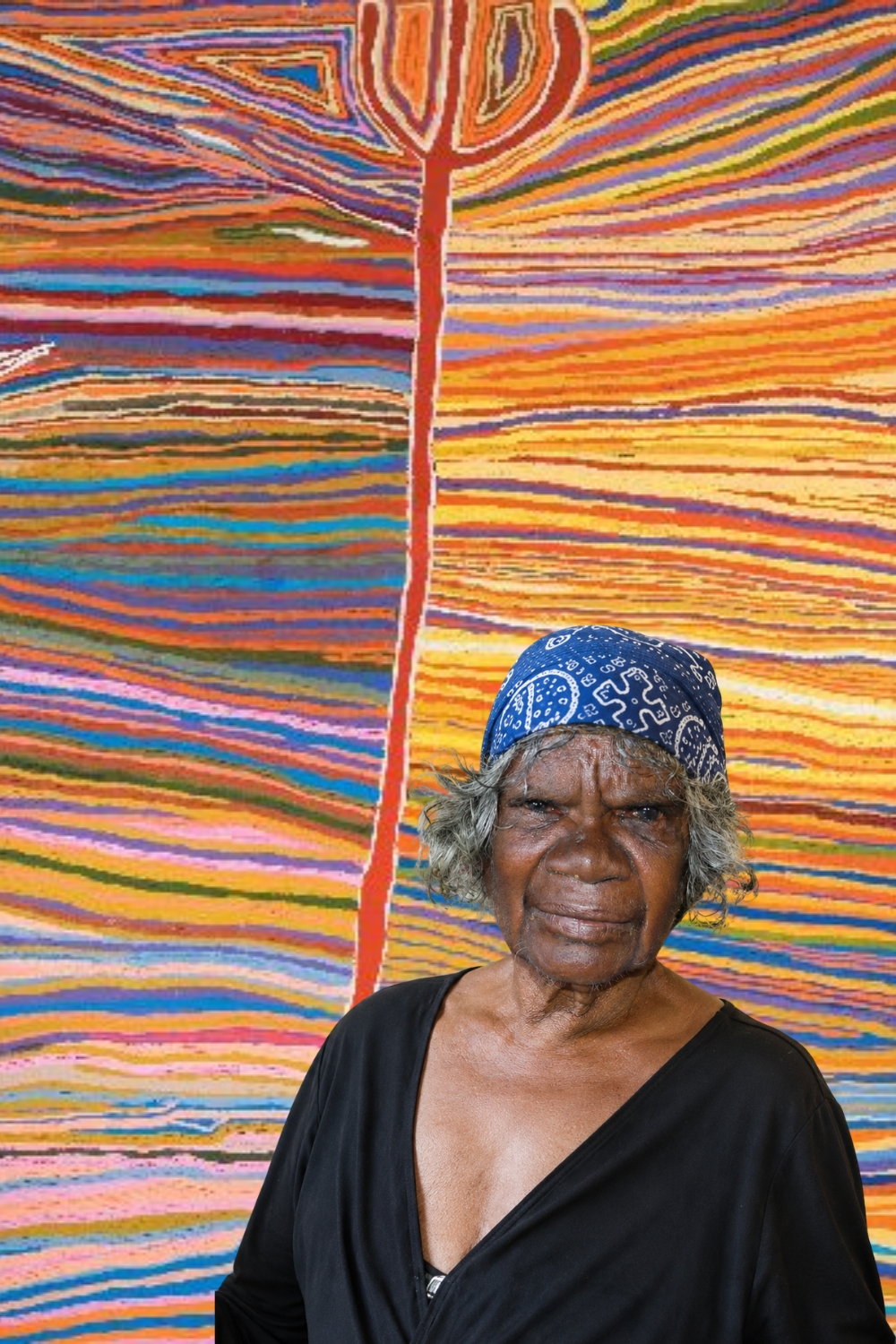 Judy Watson Napangardi – Warlpiri Artist Portrait of Judy Watson Napangardi wearing a blue patterned headscarf and black clothing, standing in front of a brightly coloured painting of flowing horizontal lines and a central red form.