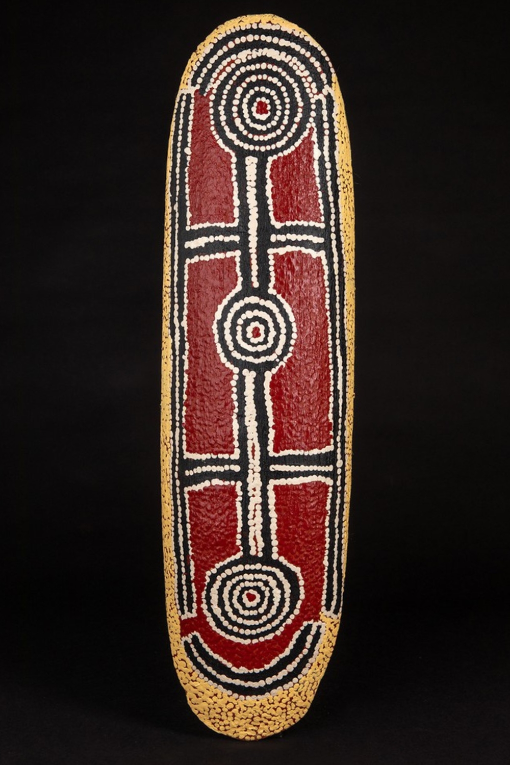 Long oval wooden shield painted by Paddy Japaljarri Stewart with three concentric circular forms connected by parallel white dotted lines, surrounded by red, black and ochre fields.