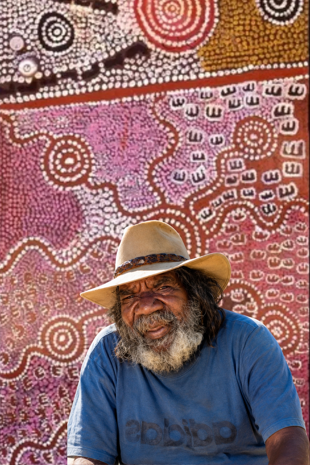 Paddy Japaljarri Stewart – Warlpiri Artist and Cultural Leader Portrait of Warlpiri artist Paddy Japaljarri Stewart wearing a broad-brimmed hat and blue shirt, seated in front of a colourful Aboriginal painting with dense dotting, concentric circles and traditional motifs