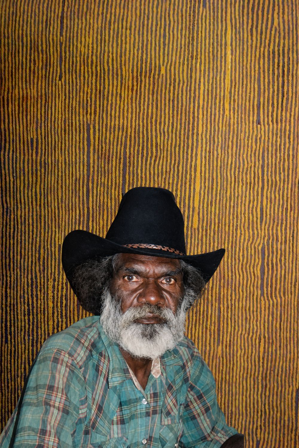 Portrait of Aboriginal artist Turkey Tolson Tjupurrula seated in front of one of his Straightening Spears paintings at Kintore, Northern Territory.