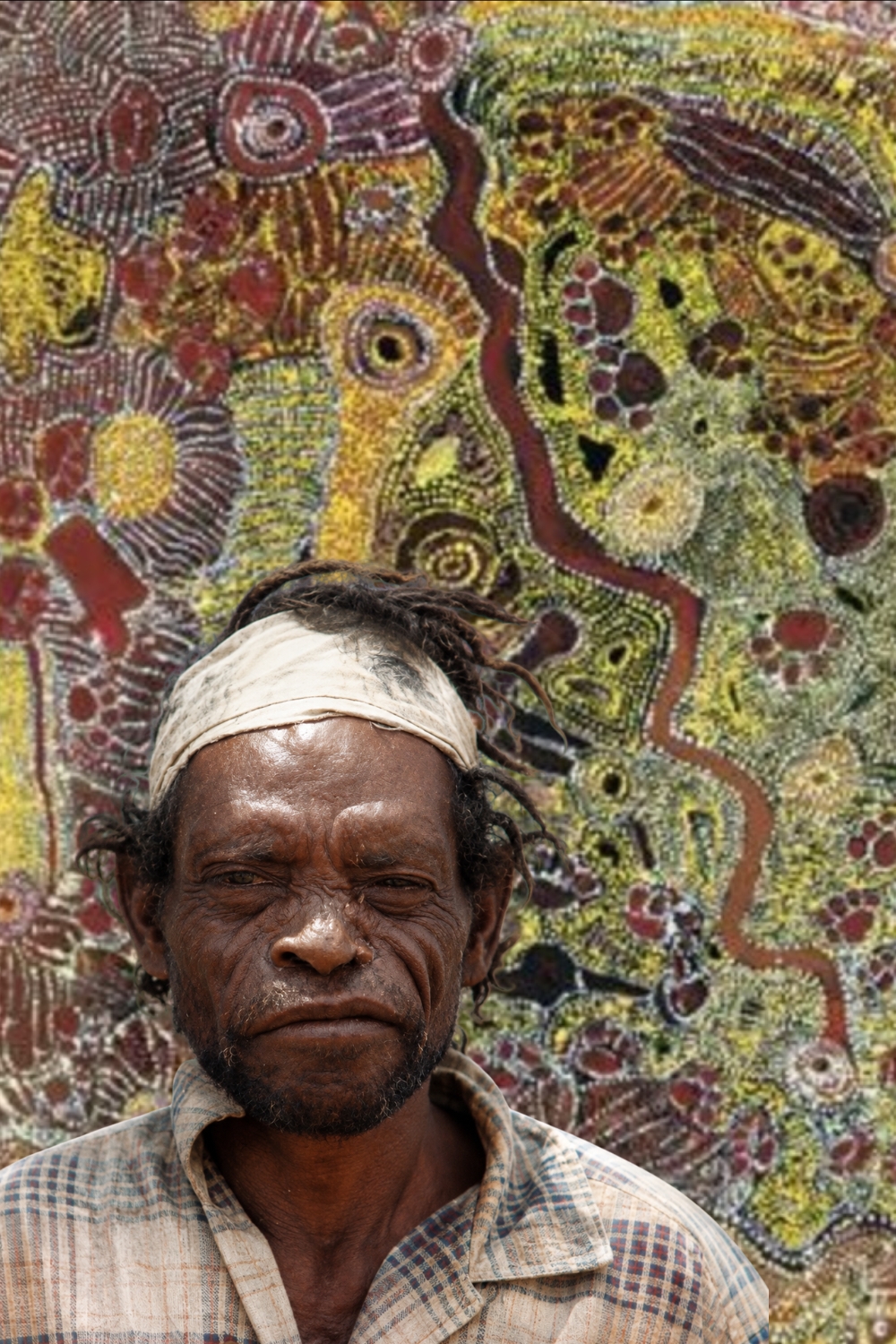 Wimmitji Tjapangarti – Balgo Aboriginal Artist Wimmitji Tjapangarti wearing a cloth headband and checked shirt, pictured in front of one of his densely dotted paintings of ancestral Country.