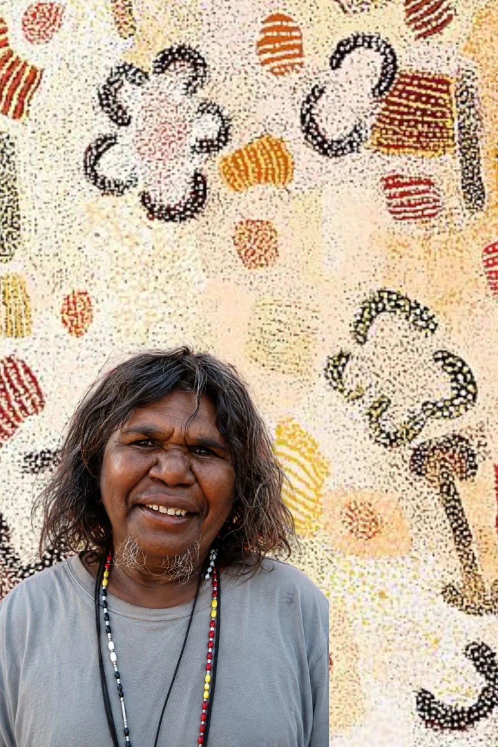 Elizabeth Nyumi Nungurrayi – Balgo Artist and Painter of Parwalla Portrait of Aboriginal artist Elizabeth Nyumi Nungurrayi standing in front of one of her pale dotted paintings of Parwalla Country.