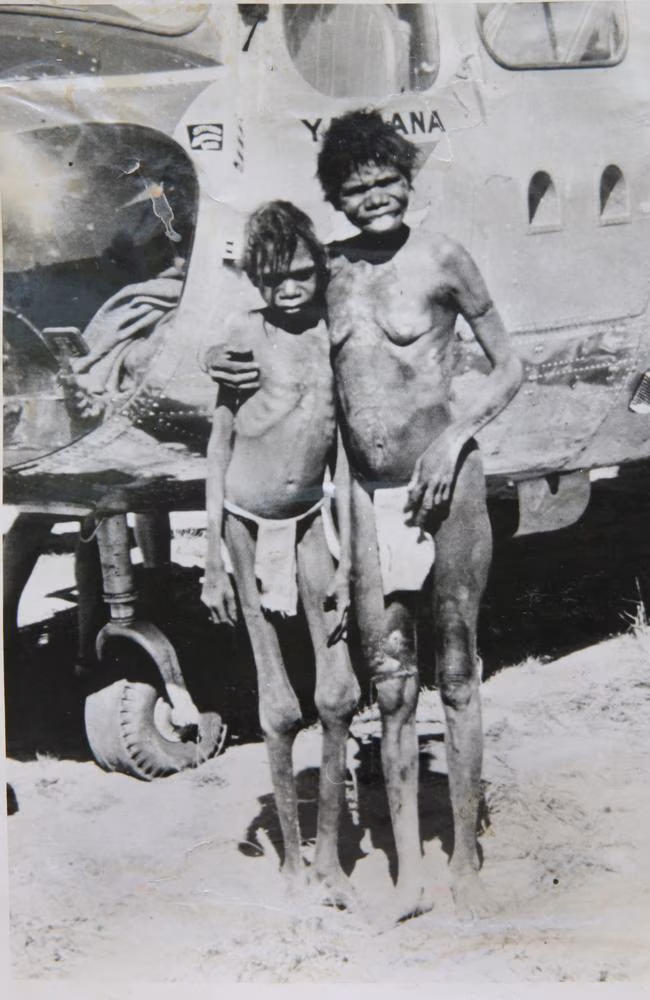 Black-and-white photograph of a young Aboriginal boy standing beside an Aboriginal woman in front of a helicopter near Natawalu on the Canning Stock Route in 1957.