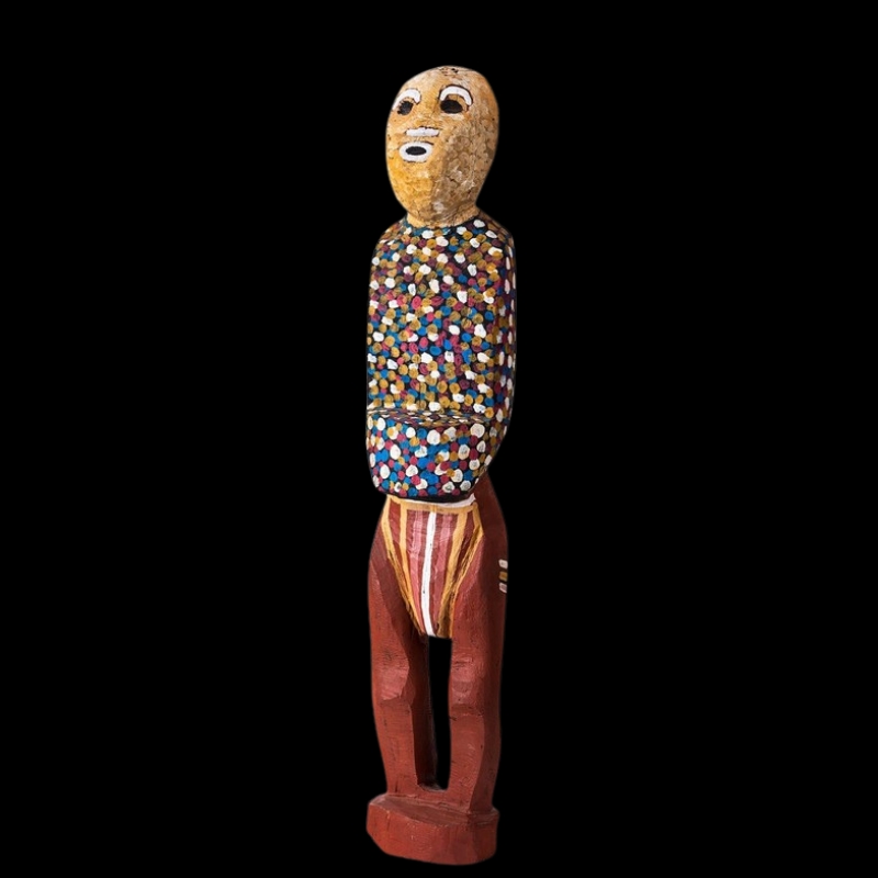 Tall carved wooden ancestral figure by Angelina Pwerle, with a multicoloured dotted torso, ochre-coloured face and red legs.
