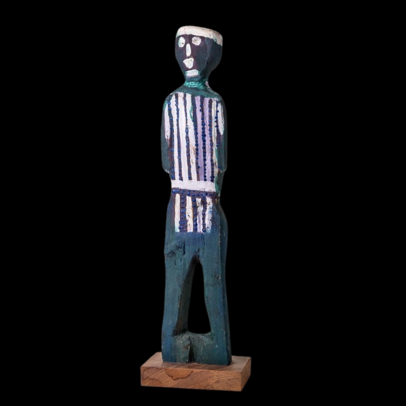 Tall carved wooden male figure by Angelina Pwerle, painted dark blue-green with white and purple vertical body markings and a simple painted face.