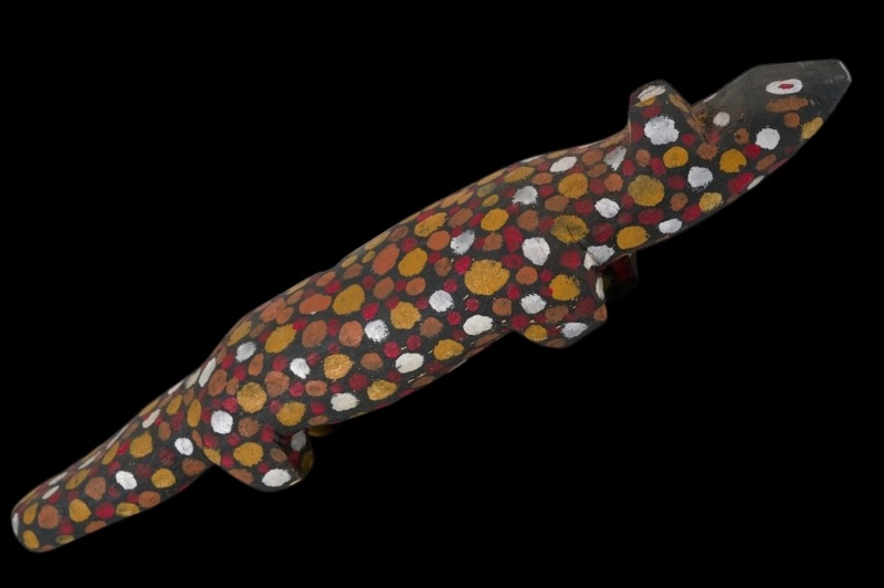 Carved wooden dog by Angelina Pwerle, painted black and covered with large red, ochre, yellow and white dots, with a long body and pointed head.