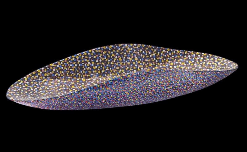 Long oval wooden coolamon by Angelina Pwerle, densely painted with small multicoloured dots in white, yellow, blue, red, pink and ochre.