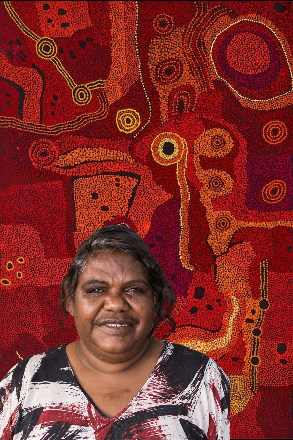 Portrait of Pitjantjatjara artist Tjungkara Ken in front of one of her richly coloured paintings of Country.