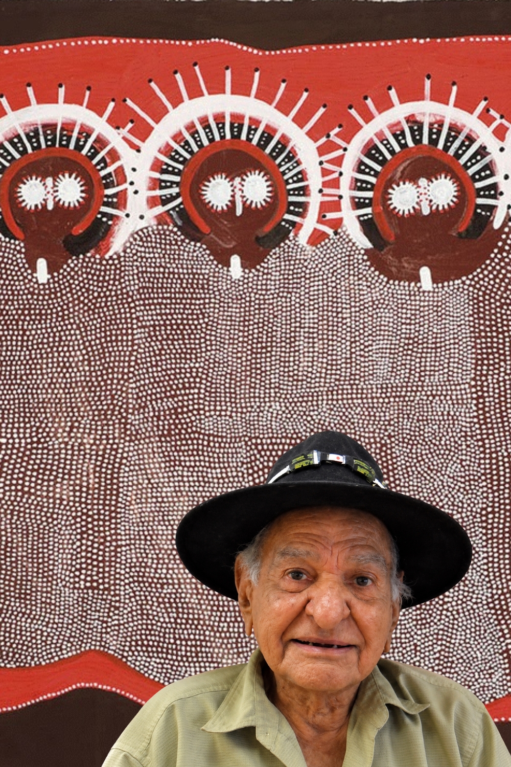 Jack Dale Mengenen – Ngarinyin Wandjina Artist Jack Dale Mengenen seated in front of a large Wandjina painting with three ancestral figures in red, white and brown.