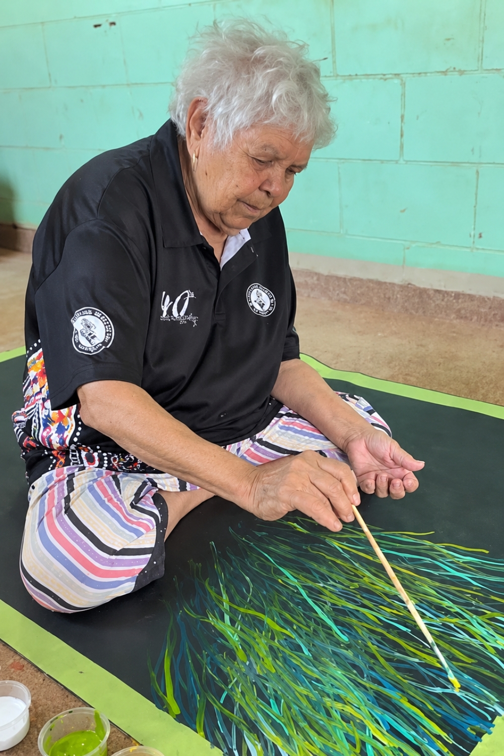 Barbara Weir painting a Grass Seed work at Utopia.  Barbara Weir seated on the floor painting with a long brush. In front of her is a dark canvas covered with overlapping green, blue and yellow linear brush marks.