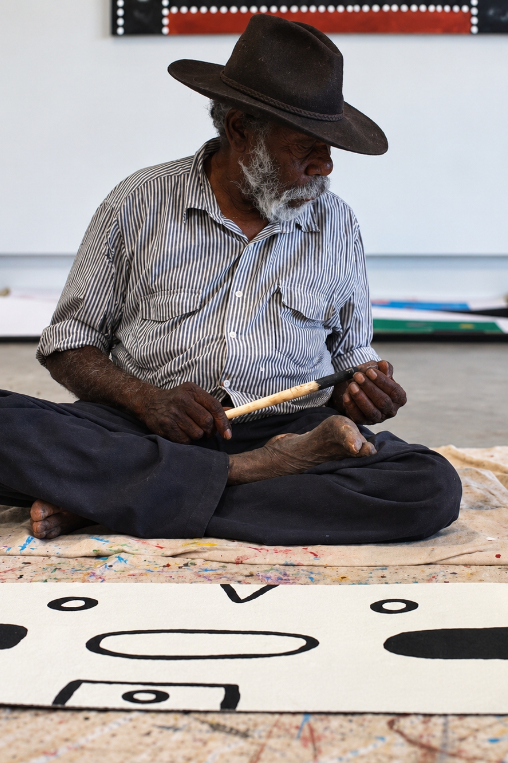 Rammey Ramsey seated cross-legged on a paint-covered studio floor, wearing a broad-brimmed hat and striped shirt while holding a paintbrush. A partly completed painting with bold black geometric forms lies on the floor in front of him.