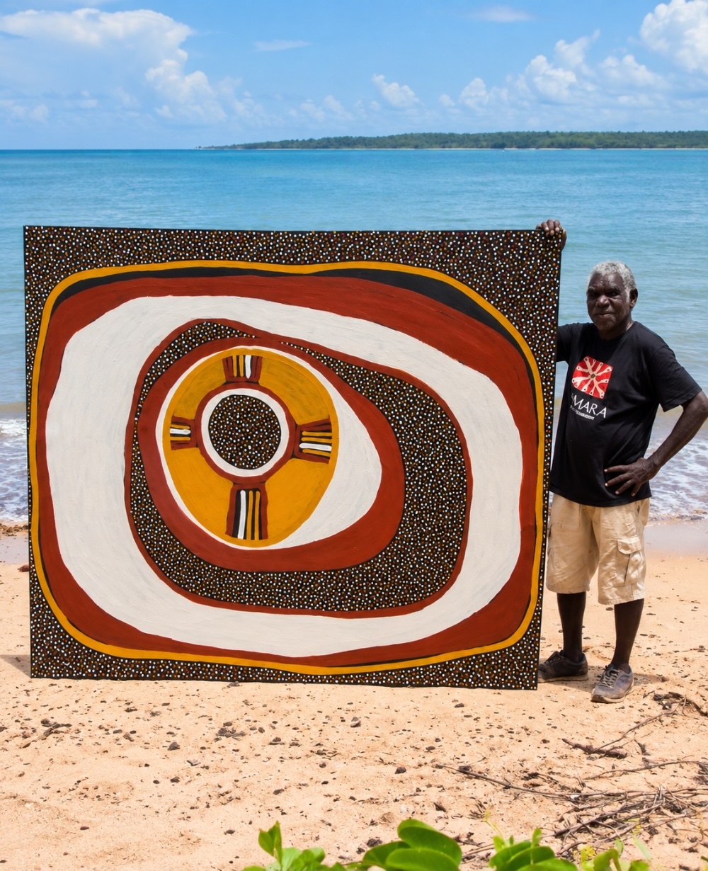 Timothy Cook standing on a beach beside a large painting with broad white, red and ochre concentric forms surrounding black fields densely covered with small dots.