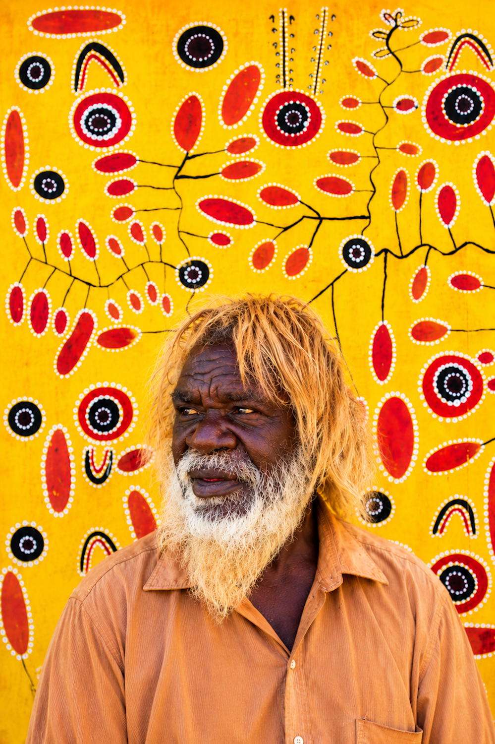 Billy Stockman Tjapaltjarri – Papunya Artist Billy Stockman Tjapaltjarri, pioneering Papunya Aboriginal artist, pictured against a colourful Western Desert painting with traditional dot designs.