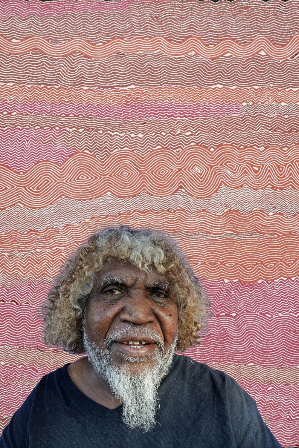 Portrait of Pintupi artist Bobby West Tjupurrula in front of a painting featuring intricate white zigzag and undulating lines on a red background.