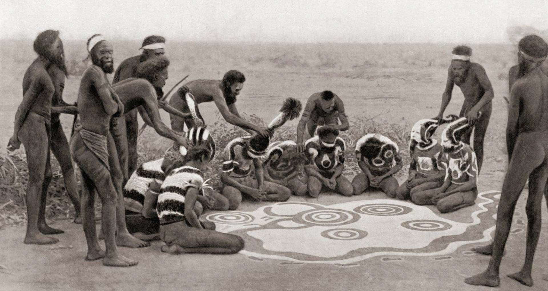 Historical photograph of Aboriginal men in Central Australia creating a ceremonial ground painting with concentric circles and curved lines, surrounded by participants wearing body paint.