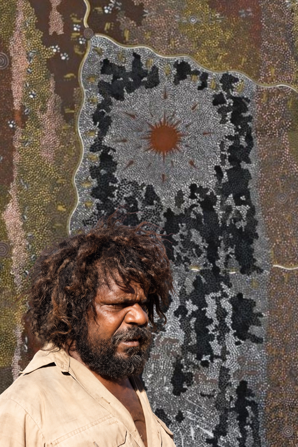 Portrait of Clifford Possum Tjapaltjarri wearing a brown shirt, against a Western Desert painting with intricate dotting, concentric circles and ceremonial designs.