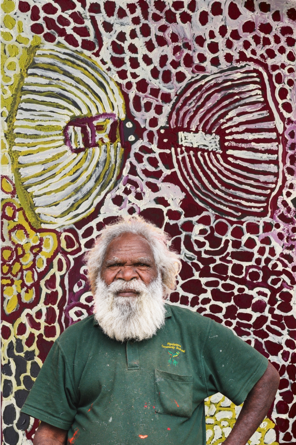 Harry Tjutjuna, Pitjantjatjara artist and traditional healer, standing before a colourful painting featuring radiating striped forms and circular patterns.