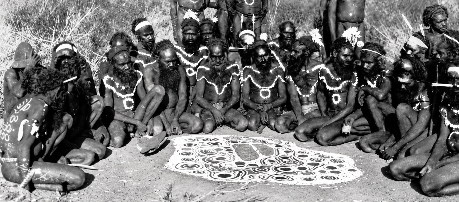 Historical photograph of Arrernte men with ceremonial body painting gathered around the Emu Ilpintira ground painting, created during an Intichiuma ceremony.