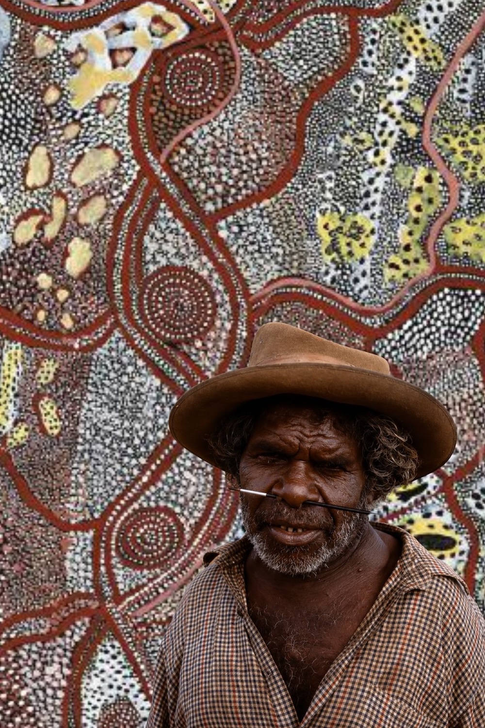 Portrait of Johnny Warangkula Tjupurrula wearing a brown hat, with a Western Desert dot painting featuring Water Dreaming imagery, concentric circles and winding pathways in the background